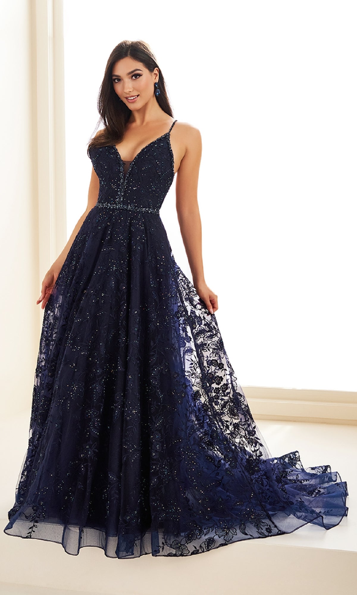 A woman is wearing the Ellie Wilde EW36088 long prom dress, featuring a deep navy blue color, thin straps, fitted bodice, and a flared embroidered tulle skirt with a small train.